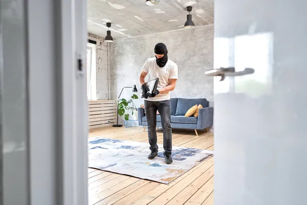 Tjuv med svart balaklava stjäla moderna dyra laptop från lägenheten. Maskerat ansikte. Man inbrottstjuv stjäla tv från huset — Stockfoto