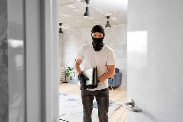 Tjuv med svart balaklava stjäla moderna dyra laptop från lägenheten. Maskerat ansikte. Man inbrottstjuv stjäla tv från huset — Stockfoto