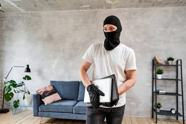 Tjuv med svart balaklava stjäla moderna dyra laptop från lägenheten. Maskerat ansikte. Man inbrottstjuv stjäla tv från huset — Stockfoto