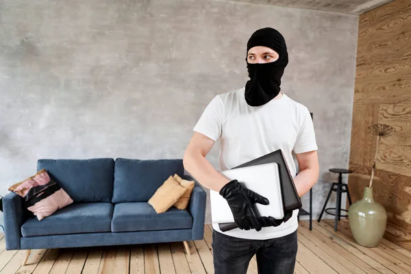 Tjuv med svart balaklava stjäla moderna dyra laptop från lägenheten. Maskerat ansikte. Man inbrottstjuv stjäla tv från huset — Stockfoto