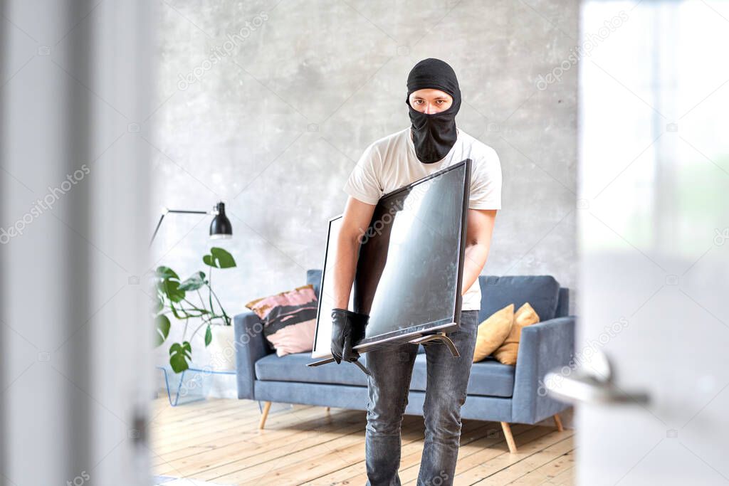 Thief with black balaclava stealing modern expensive television. Masked face. Man burglar stealing tv set from house