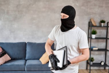 Siyah kar maskeli bir hırsız dairesinden modern ve pahalı bir laptop çalıyor. Maskeli yüz. Hırsız evden televizyon çalıyor.