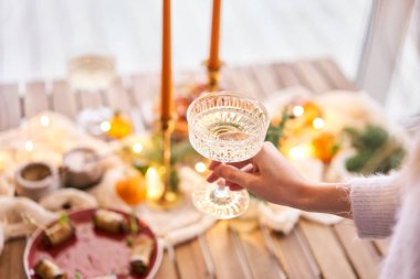 Ön planda bir bardak şampanyayla. Küçük bir masa aperatifler, bruschetalar ve kanepelerle servis edilir. Şampanya bardakları, mumlar ve Noel ağacı olan süslü bir yemek masası.