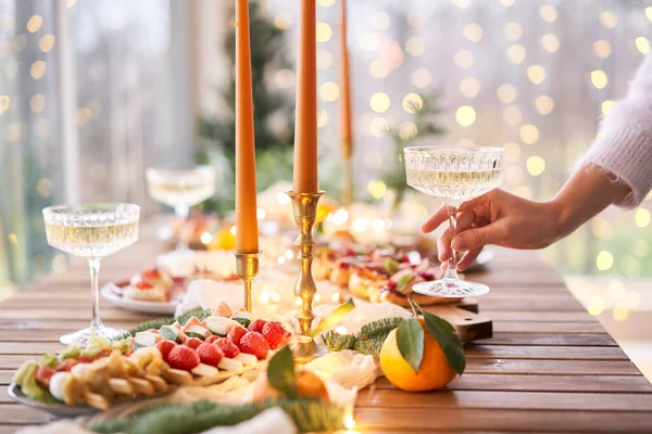 Ruku se sklenicí šampaňského v popředí. Malý stolek je servírován s občerstvením, bruschettami a konvicemi. Dekorovaný jídelní stůl se sklenicemi na šampaňské, svíčkami a vánočním stromečkem — Stock fotografie