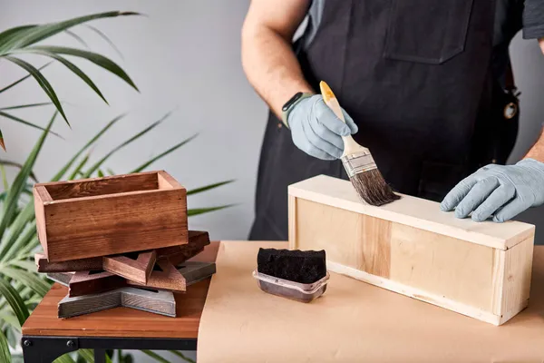 Hombre carpintero barnizado caja de madera con cepillo en su pequeño negocio. En su trabajo, ¿utiliza manchas o conservantes de madera para mostrar el patrón de madera. — Foto de Stock