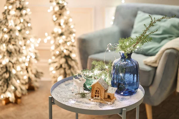 Díszített asztal a karácsonyi belső térben. Karácsonyfa. Közelkép egy kis ház, kúp, koszorú, és más karácsonyfa játékok. fény a koszorúból — Stock Fotó