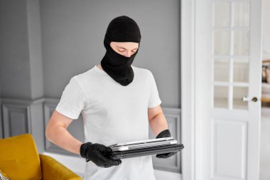 Siyah kar maskeli bir hırsız dairesinden modern ve pahalı bir laptop çalıyor. Maskeli yüz. Hırsız evden televizyon çalıyor.