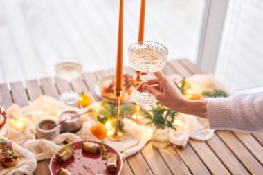 Ön planda bir bardak şampanyayla. Küçük bir masa aperatifler, bruschetalar ve kanepelerle servis edilir. Şampanya bardakları, mumlar ve Noel ağacı olan süslü bir yemek masası.