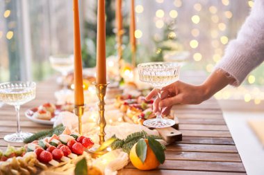 Ön planda bir bardak şampanyayla. Küçük bir masa aperatifler, bruschetalar ve kanepelerle servis edilir. Şampanya bardakları, mumlar ve Noel ağacı olan süslü bir yemek masası.