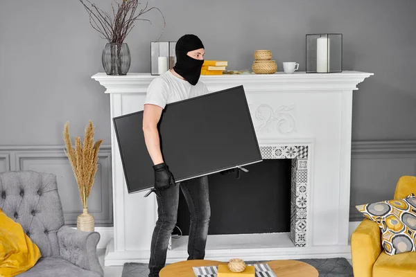 Ladrão com balaclava preta roubando televisão moderna cara. Cara mascarada. Homem ladrão roubar tv set de casa — Fotografia de Stock