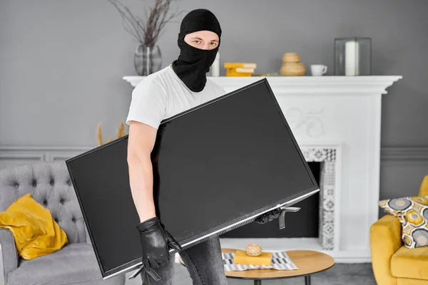 用黑色巴拉克拉瓦偷窃现代昂贵电视机的贼。蒙面脸男子窃贼从家里偷电视机 — 图库照片