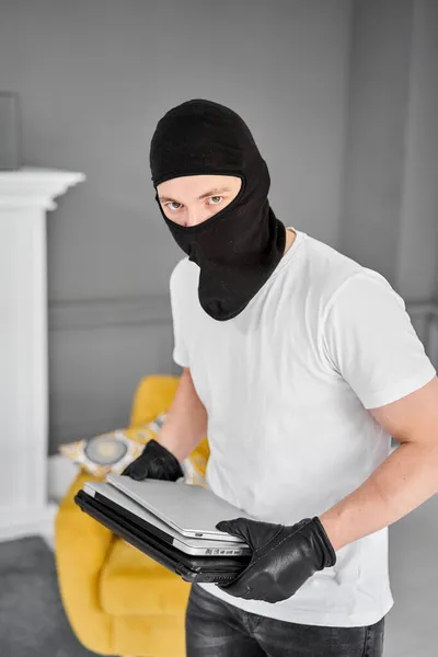 Siyah kar maskeli bir hırsız dairesinden modern ve pahalı bir laptop çalıyor. Maskeli yüz. Hırsız evden televizyon çalıyor. — Stok fotoğraf