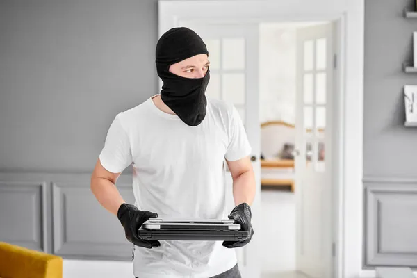 Ladrão com balaclava preta roubando laptop moderno caro do apartamento. Cara mascarada. Homem ladrão roubar tv set de casa — Fotografia de Stock