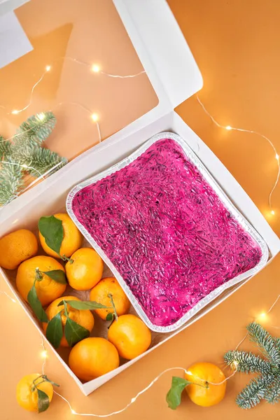 Russian traditional salad, dressed herring under fur coat in take way box. Menu food for delivery in the Coronavirus Pandemic. Tangerines and branches of the Christmas tree. Russian national cuisine.