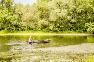 A sport fisherman rides on a pond in a wooden boat with the help of an oar.