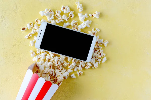 Karton bardakta patlamış mısır ve sarı arka planda cep telefonu. Boş zaman konsepti, film zamanı, dinlenme. Kopyalama alanı olan üst görünüm.