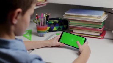 Çocuk elinde yeşil ekranlı akıllı bir telefon tutuyor, ekranda kayıyor. Genç çocuk masada oturuyor ve uzaktan eğitim için izole edilmiş arka plan ve model şablonuyla ekrana bakıyor.