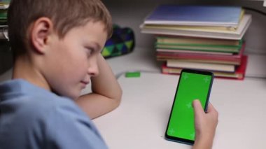 Çocuk elinde yeşil ekranlı akıllı bir telefon tutuyor, ekranda kayıyor. Genç çocuk masada oturuyor ve uzaktan eğitim için izole edilmiş arka plan ve model şablonuyla ekrana bakıyor.