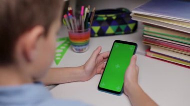 Çocuk elinde yeşil ekranlı akıllı bir telefon tutuyor, ekranda kayıyor. Genç çocuk masada oturuyor ve uzaktan eğitim için izole edilmiş arka plan ve model şablonuyla ekrana bakıyor.