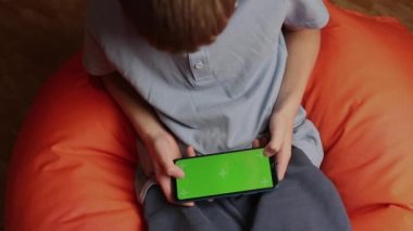 Çocuk turuncu puf koltuğa oturur ve yeşil ekranlı akıllı telefonu tutarak cep telefonuyla oyunlar oynar.