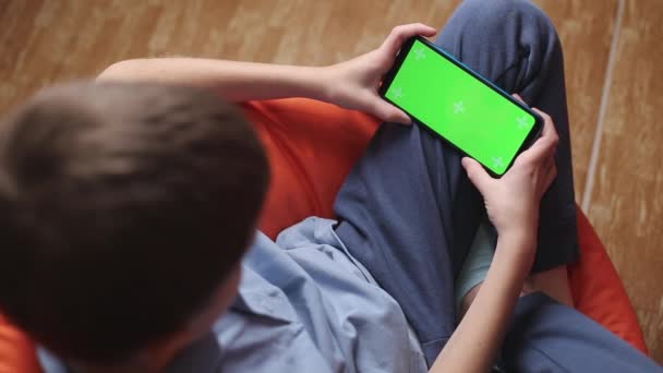 Çocuk turuncu puf koltuğa oturur ve yeşil ekranlı akıllı telefonu tutarak cep telefonuyla oyunlar oynar. — Stok video
