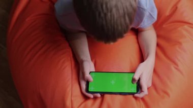 Çocuk turuncu puf koltukta uzanıyor ve cep telefonuyla oyunlar oynuyor, elinde yeşil ekranlı akıllı telefon var.