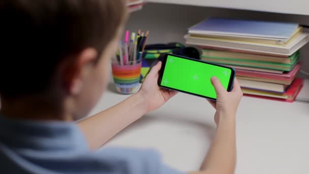 집에 녹색 화면 이 있는 스마트폰을 들고 있는 남자 아이가 터치 스크린을 스크롤합니다. 어린 소년은 테이블에 앉아서 외진 교육을 위한 모의 템플릿 과고립된 배경을 가지고 전시를 보고 있습니다. — 비디오