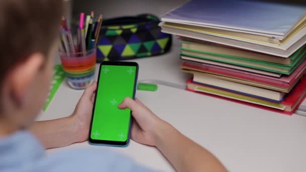 Niño sosteniendo un teléfono inteligente con una pantalla verde en casa, desplazándose pantalla táctil. Niño se sienta en la mesa y mirando a la pantalla con plantilla de maqueta y fondo aislado para la educación remota — Vídeo de stock
