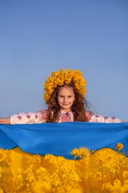 Ukrainian child girl in embroidered shirt and yellow wreath in field of yellow flowers against blue sky. Ukraine blue-yellow flag in hands of little Ukrainian girl. Independence Flag Constitution day.