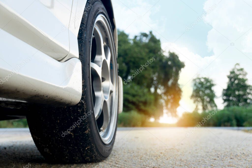 Close-up skirt of Beside white car. View of the front suspension vehicle that sees the wheels and tires on a dirt road. Aim at the destination in front of green trees and orange sunlight.