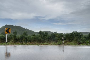 Tayland 'daki ıslak asfalt yolunun yatay görüntüsü. Yağmurlu zaman ortamı. Cassava çiftliğinin ve yeşil dağların geçmişi. Mavi gökyüzü ve kara bulutların altında. kıvrımlı yol işareti ile.