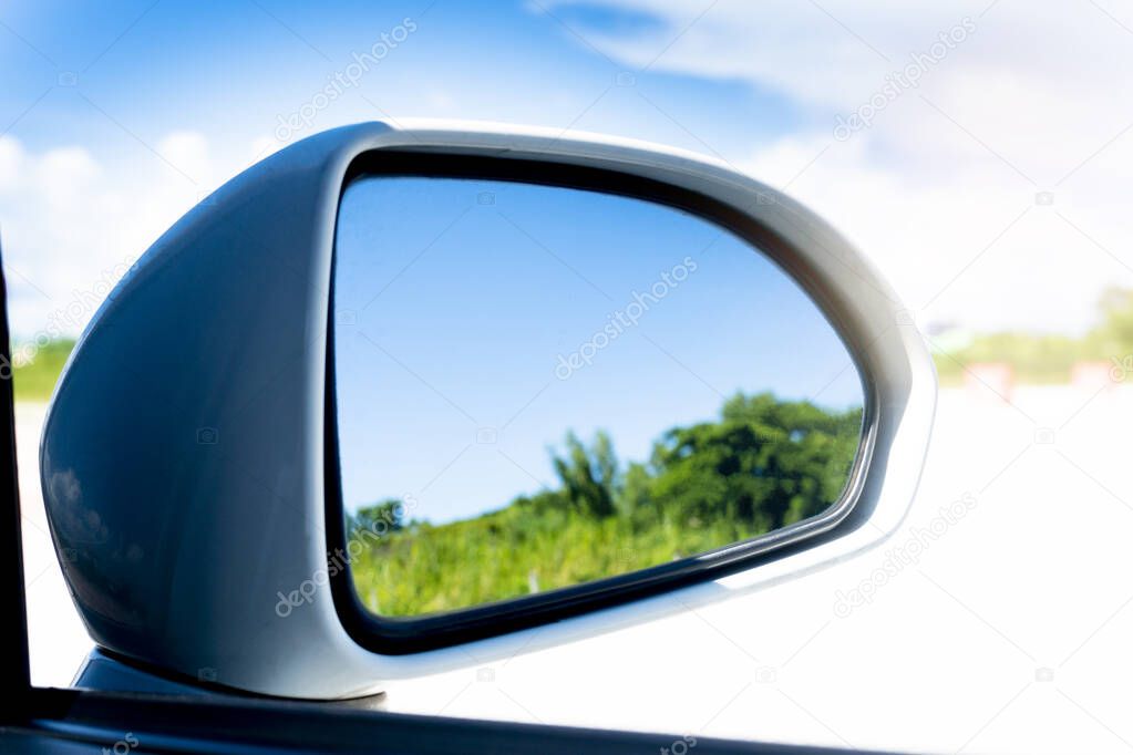 Mirror wing of white car with reflection of green trees blur in mirror. Under sky was bright and turquoise. Bright outdoor spaces.