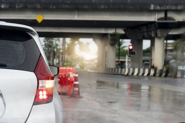 Rear side of white car with turn signal wet with rain. Concrete road surface wet with rainy. Parked at a red traffic light under a leveled bridge. Blurred water droplets from the road surface.