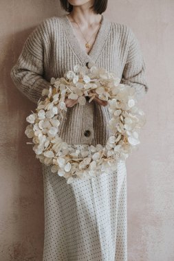 Bej renkli kazaklı ve etekli kadın, nötr tozlu pembe duvarın önünde altın ay yapraklarından bir Noel çelengi tutuyor. Zarif estetik minimalist moda geçmişi