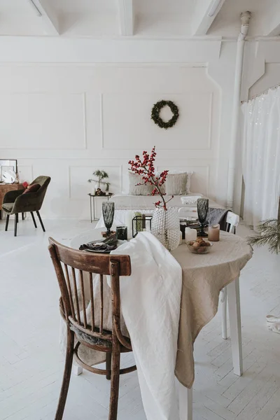 Elegant aesthetic Christmas dinner serving concept. Bright living room interior with chair, table with beige linen table cloth, berries bouquet. Christmas celebration evening
