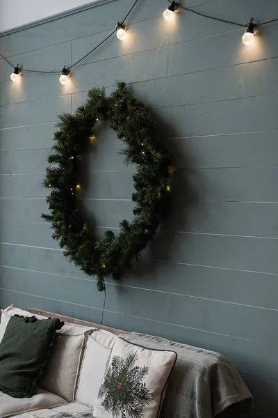 家庭内部舒适舒适的沙发 灿烂的花环灯 冷杉圣诞花环挂在尘土飞扬的蓝色墙壁上 斯堪的纳维亚风格的圣诞装饰 — 图库照片