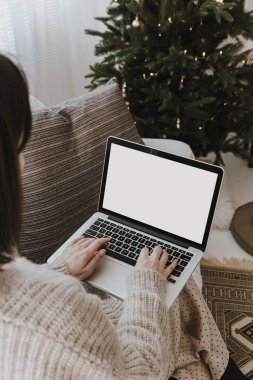 Genç bir kadın dizüstü bilgisayarda çalışıyor. Boş ekran ve fotokopi alanı var. Noel için dekore edilmiş modern ev dekorasyonu, Noel ağacı ile yeni yıl kutlaması..
