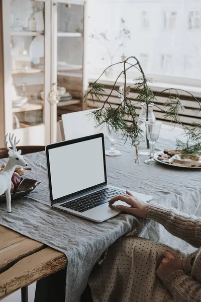 Woman work on laptop computer with blank screen on linen table cloth with fir spruce bouquet, Christmas decorations. Elegant home office working space interior design. Freelancer works at home