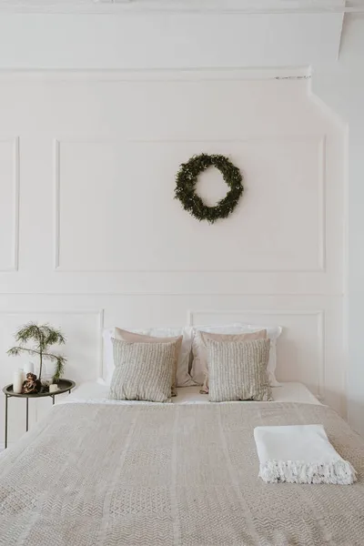 Modern bright home bedroom interior with bed, neutral bed linens, pillows, Christmas wreath made of pine needles hanging on white wall. Aesthetic living room with Christmas decorations