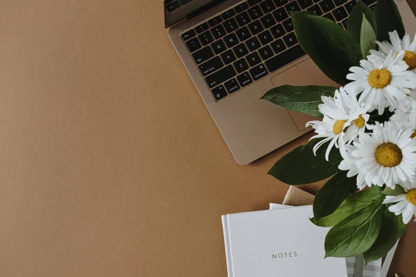 Aesthetic minimalist home office workspace desk. Laptop computer, notebook, daisy flowers bouquet on dark brown background. Flat lay, top view work, business concept