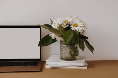 Masasında papatya çiçeği buketi olan boş ekranlı dizüstü bilgisayar. Estetik etki alanı biçimlendirilmiş çalışma alanı iç tasarım şablonu kopya alanı ile