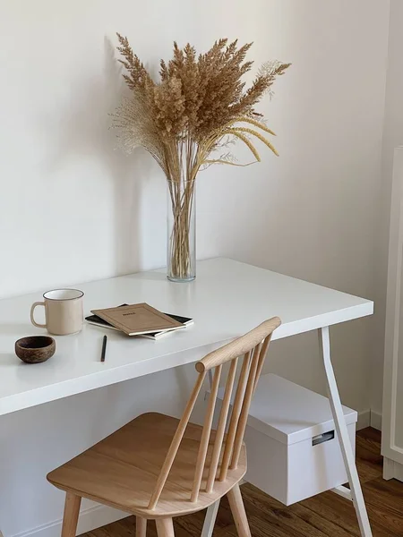 Aesthetic minimal office workspace interior design. Chair and table. Mug, notebook, pampas grass floral bouquet on white table against white wall. Girl, woman boss work at home business concept