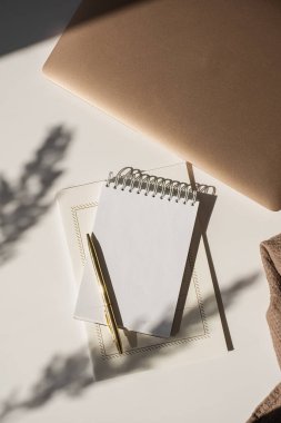 Fotokopi alanı, güneş ışığı gölgeleri ve beyaz arka planda dizüstü bilgisayarı olan boş bir kâğıt defter. Estetik lüks minimalist çalışma alanı. Düz yatış, üst görünüm
