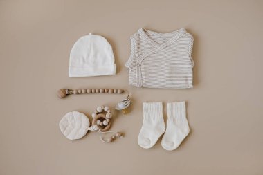 Tarafsız pastel bej arka planda zarif bebek kıyafetleri, aksesuarlar ve oyuncak kolajı. Temel vücut kıyafeti, şapka, emzik, çoraplar. Düz yatış, üst görünüm
