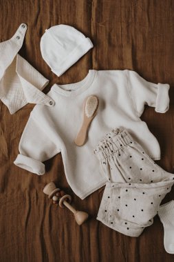 Muslin önlüğü, kazak, koşucular, fırça, çorap, şapka. Yeni doğan bebek kıyafetleri, aksesuarlar ve kahverengi keten battaniyeye dizilmiş oyuncaklar. Moda İskandinav yeni doğan temel ihtiyaçları. Düz yatış, üst görünüm