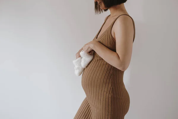 Young pregnant woman in brown dress hold white newborn socks against white wall. Elegant aesthetic minimalist pregnancy, maternity concept. Baby expecting
