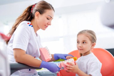 Pediatrik dişçi küçük bir kızın dişlerini nasıl düzgün fırçalayacağını gösterir. Güzel kız dişçinin ofisinde gülümsüyor. konsept bir çocuk tıbbi muayenesidir..