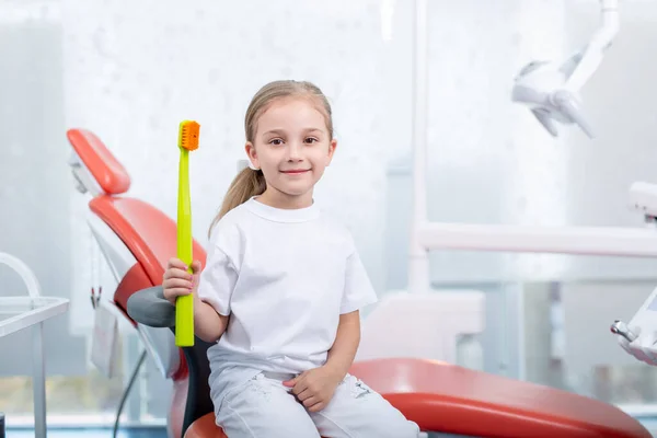 Çocuk doktorunun resepsiyonundaki beyaz elbiseli kız gülümsedi ve güldü. Küçük kız doktorun ofisinde büyük bir diş fırçası tutuyor. konsept çocuk sağlığı..