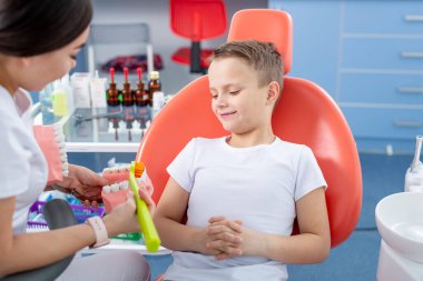 Dişi dişçi genç bir çocuğa dişlerini nasıl fırçalayacağını gösteriyor. Yakışıklı çocuk dişçinin ofisinde gülümsüyor. konsept bir çocuk tıbbi muayenesidir..