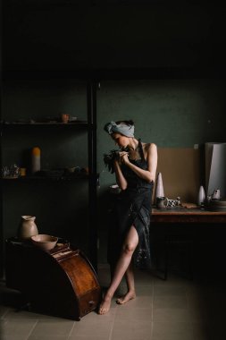 Kafasında bandaj ve çıplak vücudunda siyah bir önlükle güzel heykeltraş kız büyük pencerenin yanındaki çömlekçilik atölyesinde duruyor. konsept güzellik